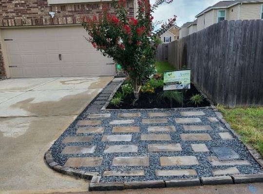 Front yard garden bed with stepping-stone pavers, a flowering tree, and a real-estate sign beside a driveway