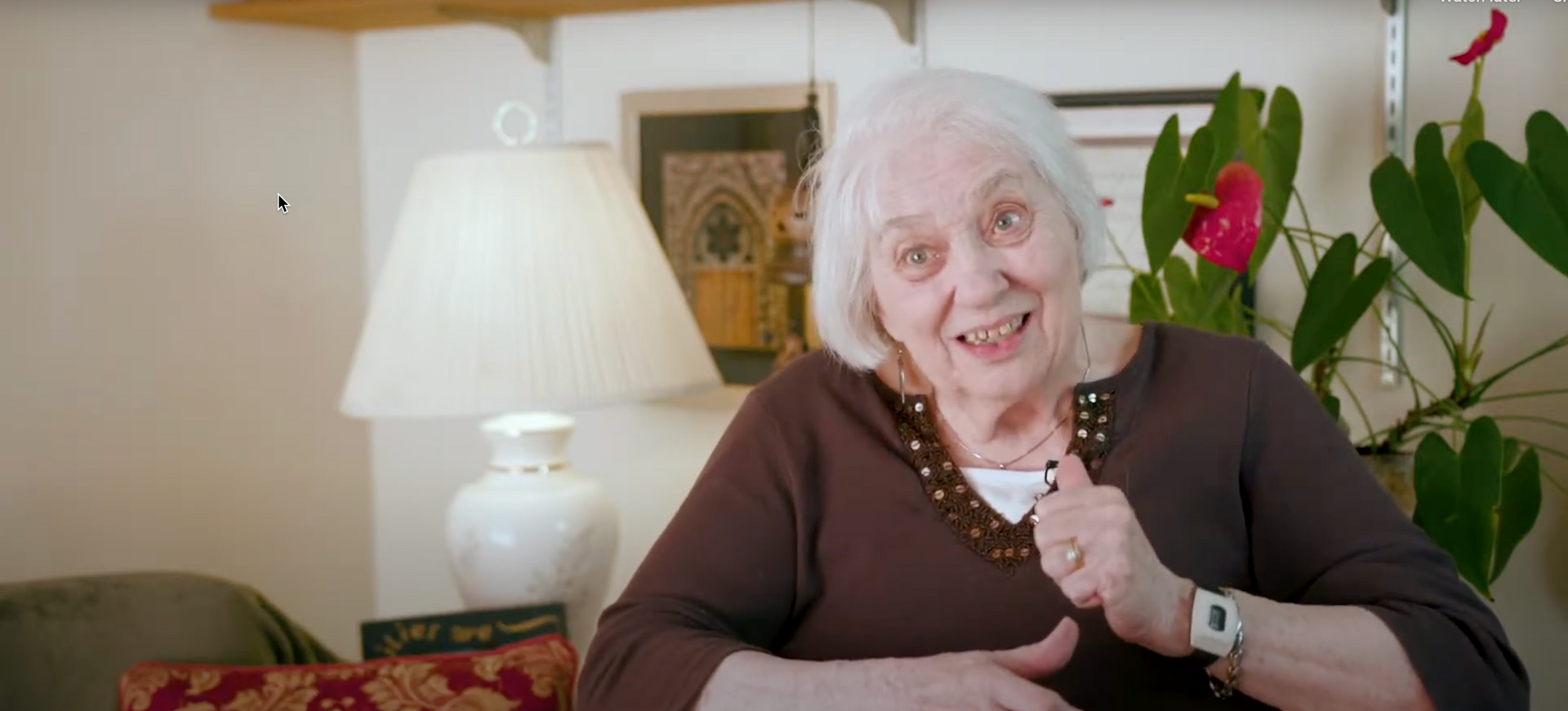 An elderly woman is sitting in a living room with a lamp.