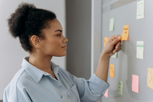 Femme plaçant un post-it sur un tableau. Chemise bleu clair, coupe afro, expression neutre.