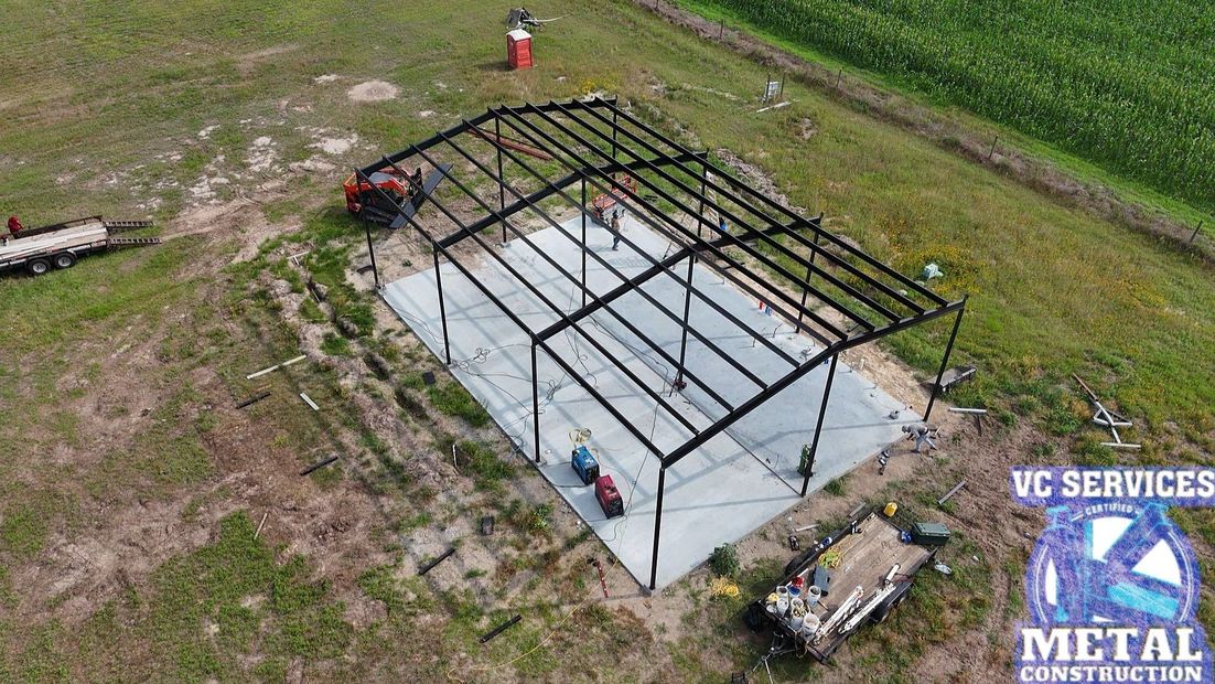 Aerial view of a steel building frame on a concrete slab with workers and equipment on a grassy site