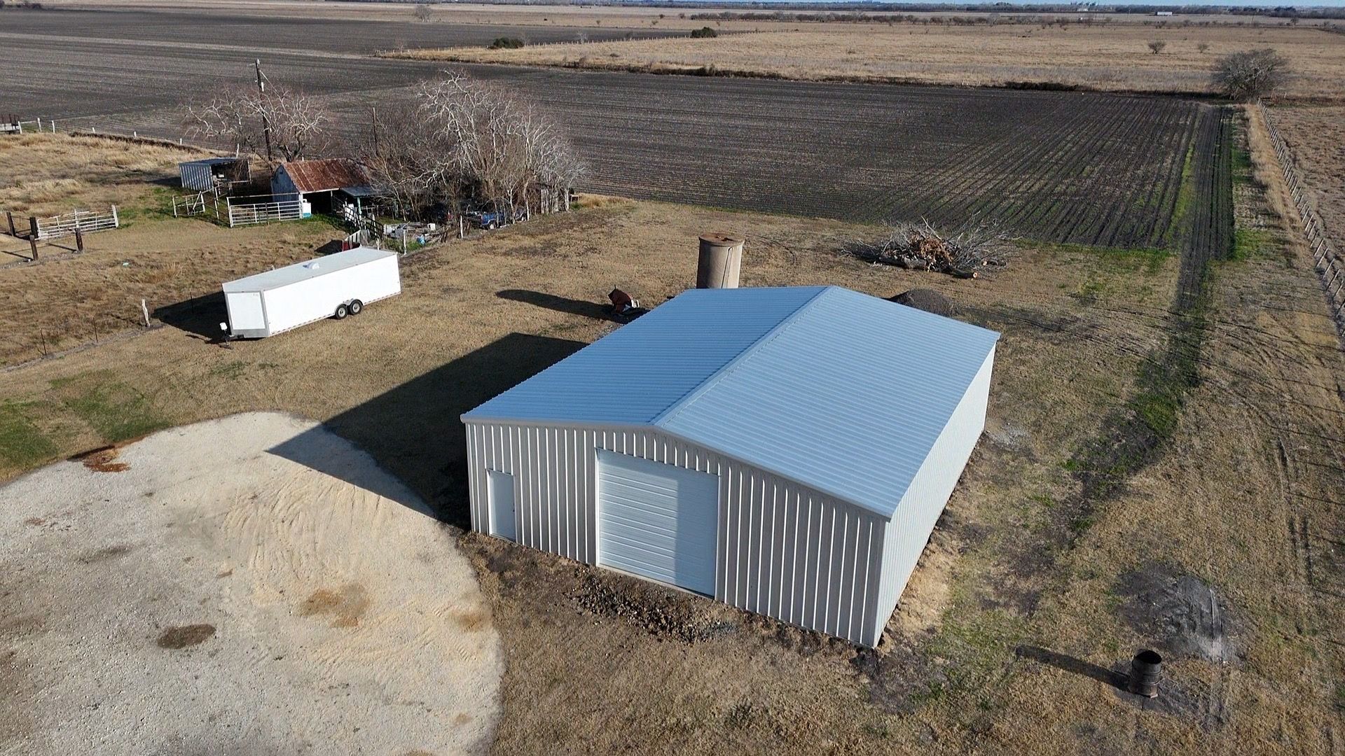 Aerial view of a metal-roofed shed in a rural field with a trailer and open land nearby