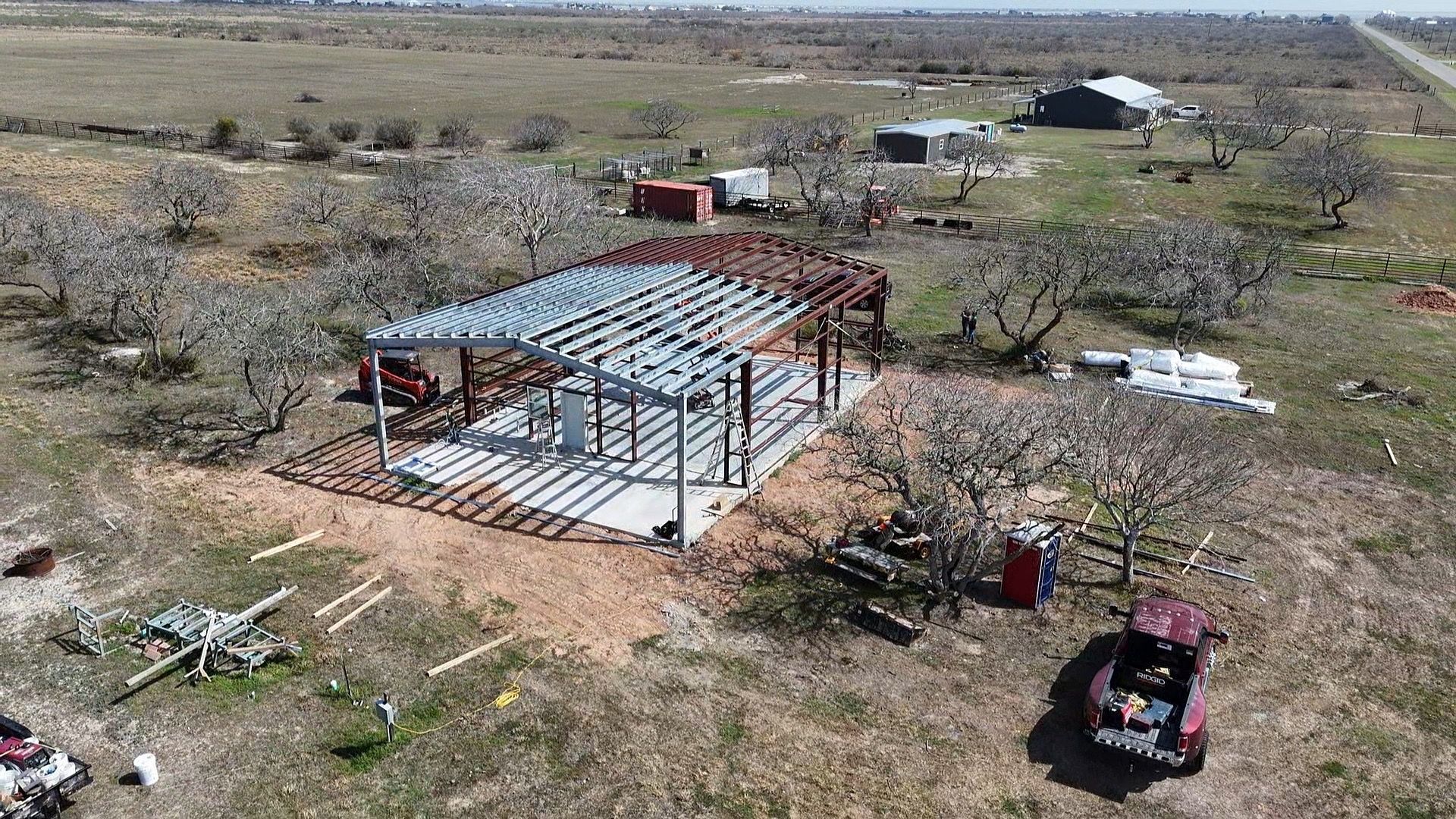 Aerial view of a partially built metal-roof structure in a dry rural field with vehicles nearby