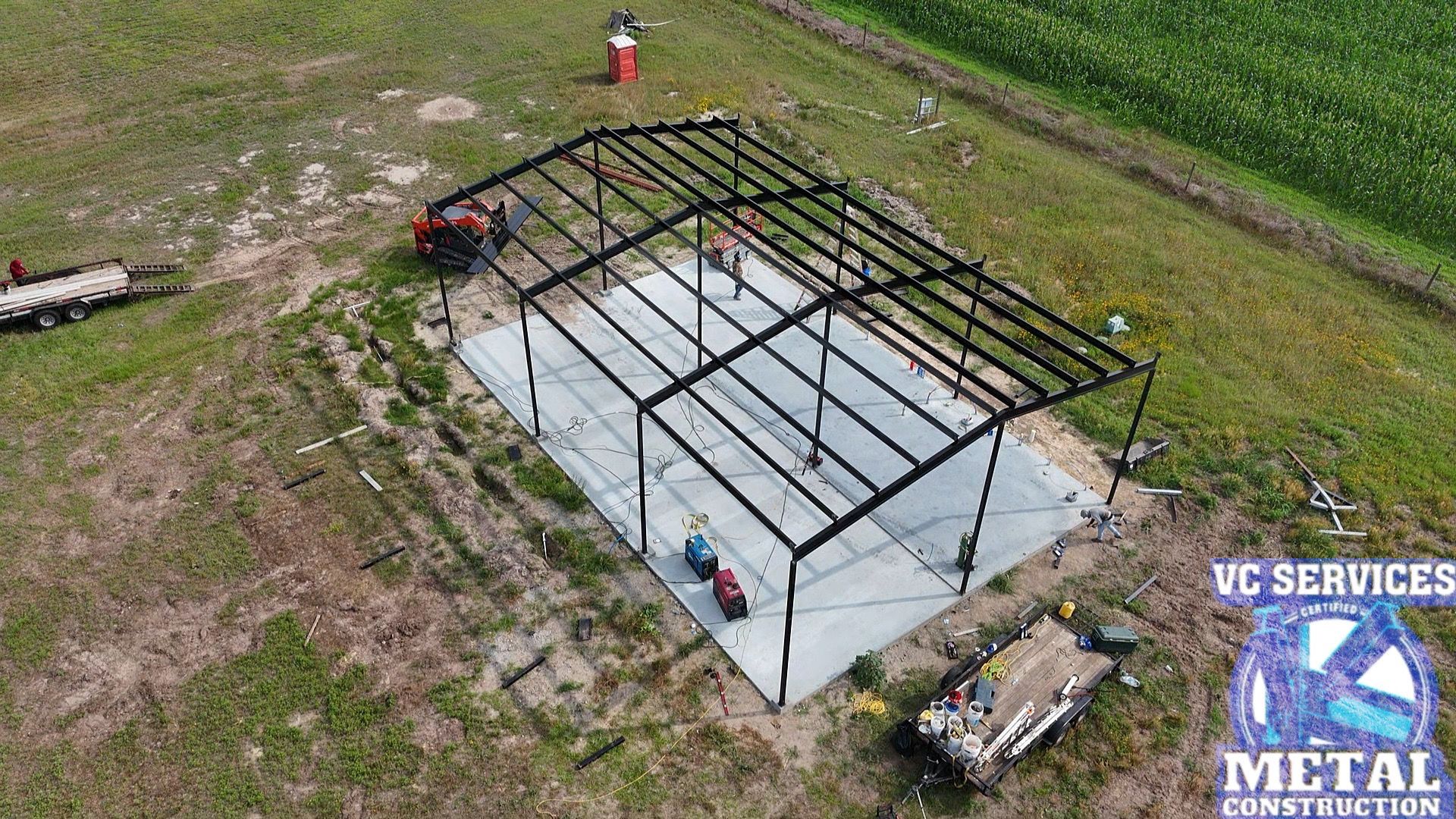 Aerial view of a steel building frame on a concrete slab with workers and materials nearby.