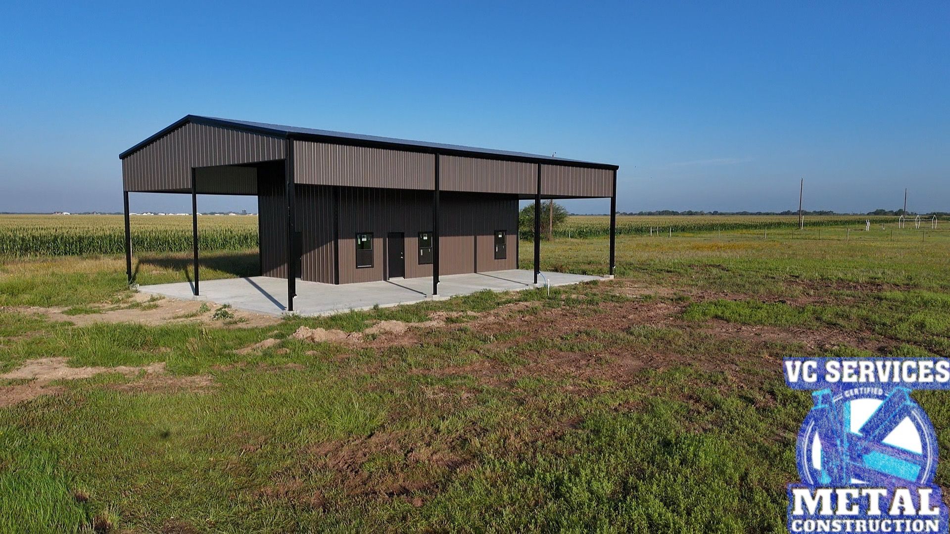 Small black metal shed in a grassy field under a clear blue sky, with VC Services logo watermark.