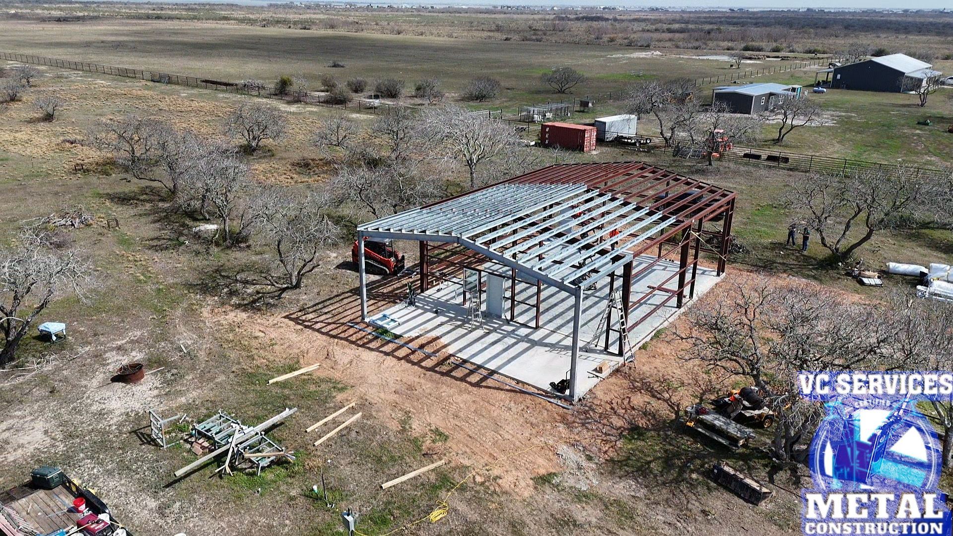 Steel-frame building under construction in a rural field, with scattered materials and a red container nearby.