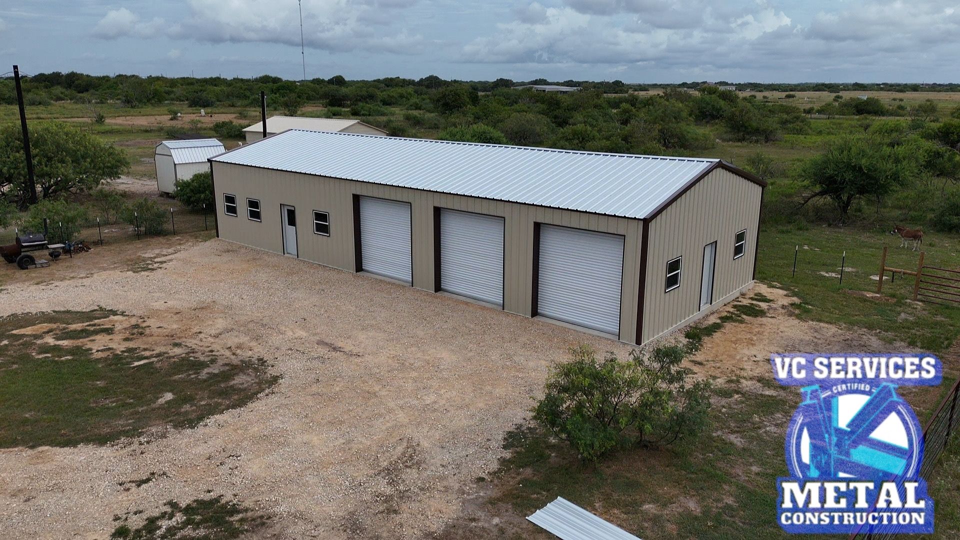 White metal service building with roll-up doors on a gravel lot in a rural field, viewed from above