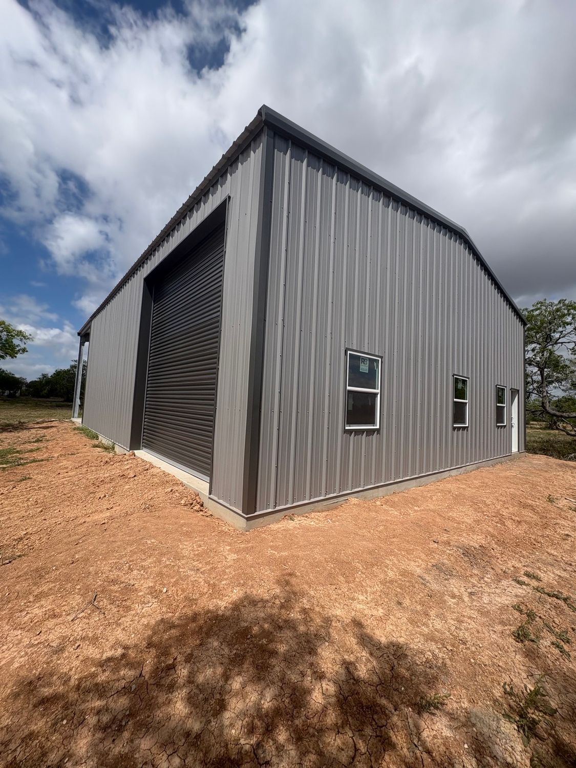 Gray metal warehouse with large roll-up door, set on a gravel lot under a cloudy sky