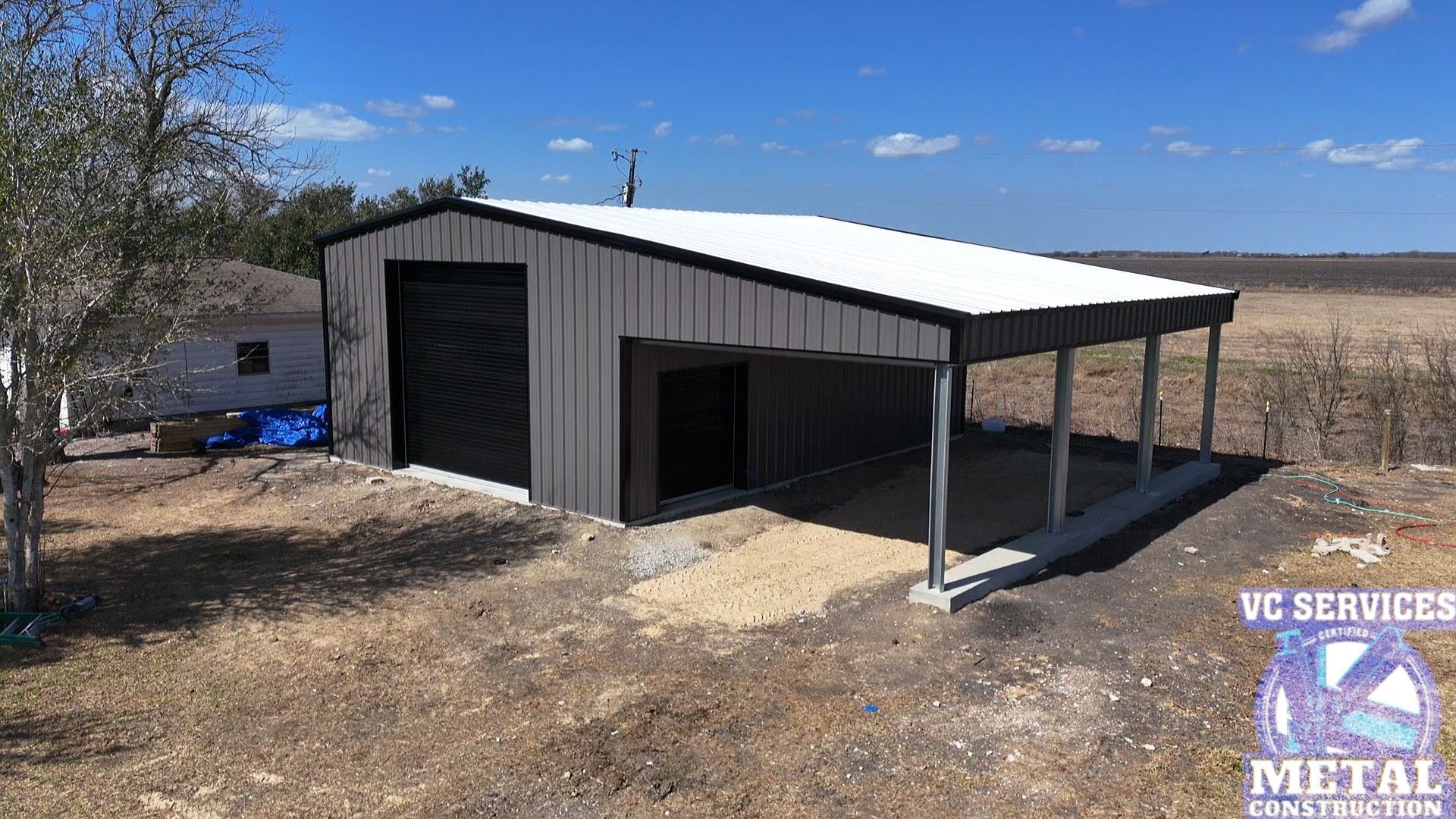 Metal-sided shed with open front porch and black garage door in a dry rural field.