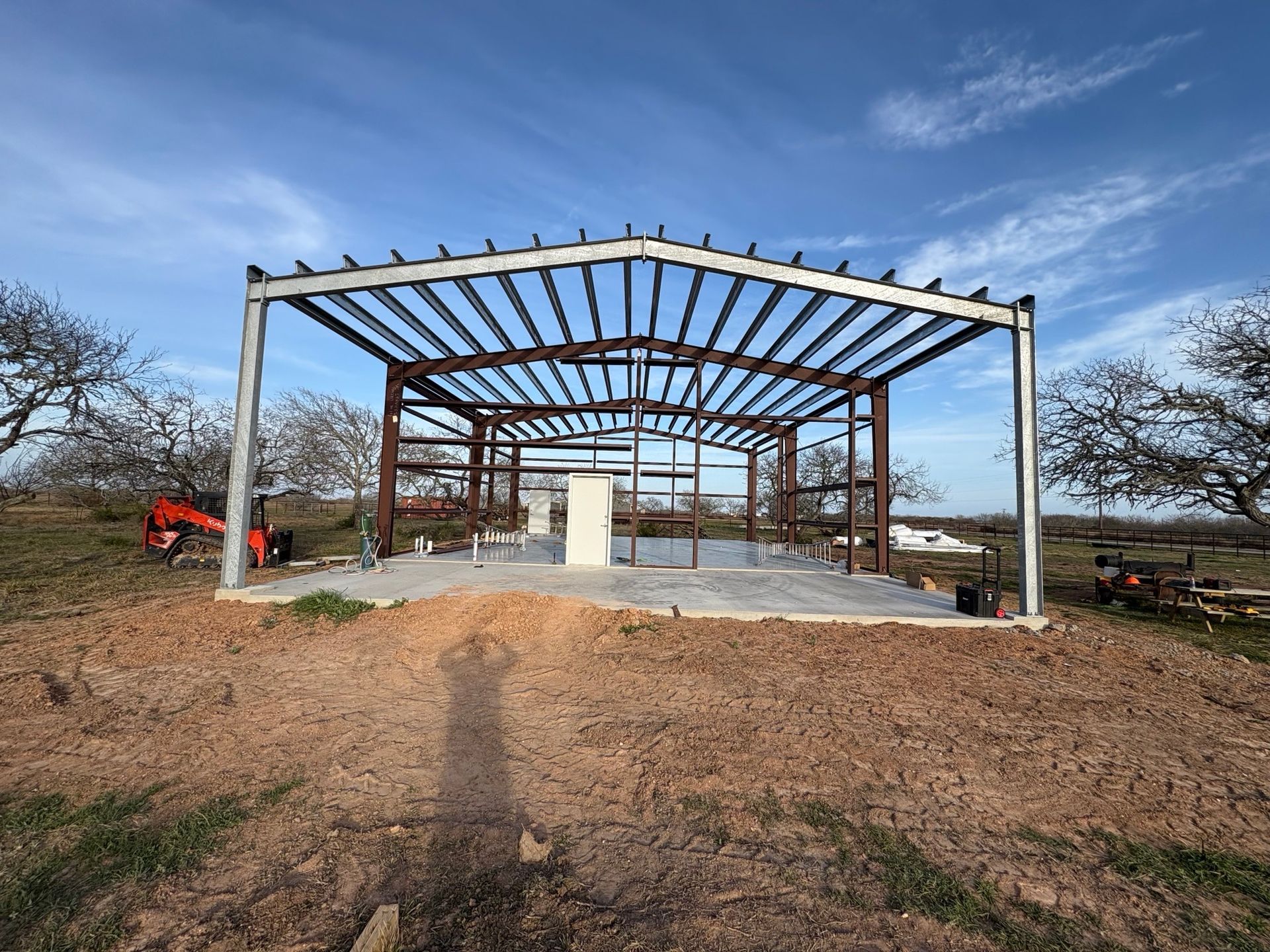 Partially built steel pavilion frame on a concrete pad in a rural field under a blue sky