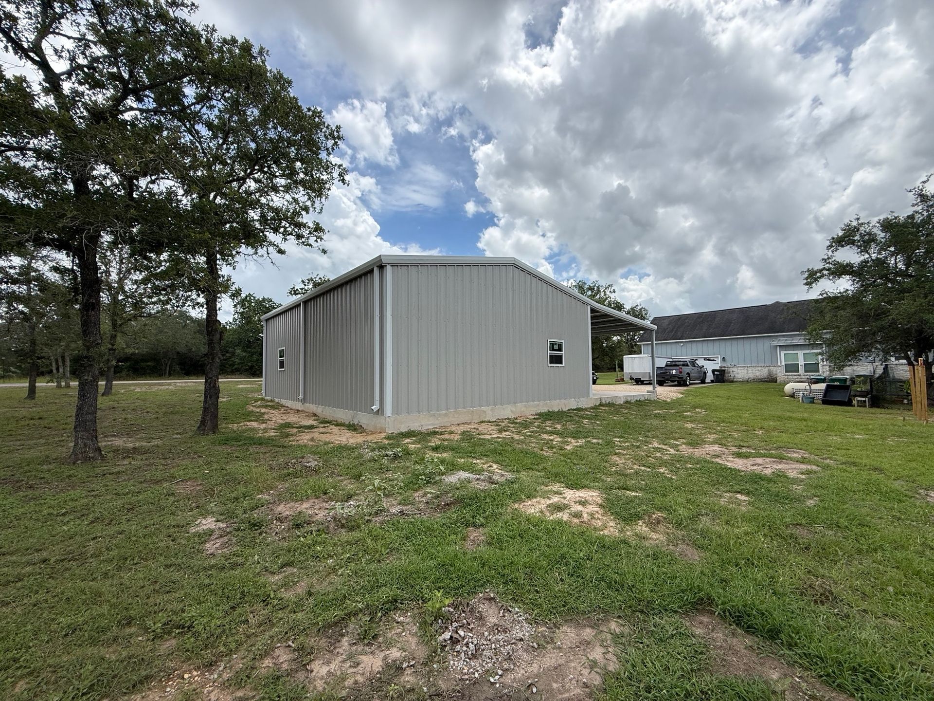 Gray metal building on a grassy lot under cloudy sky, with trees and parked vehicles nearby