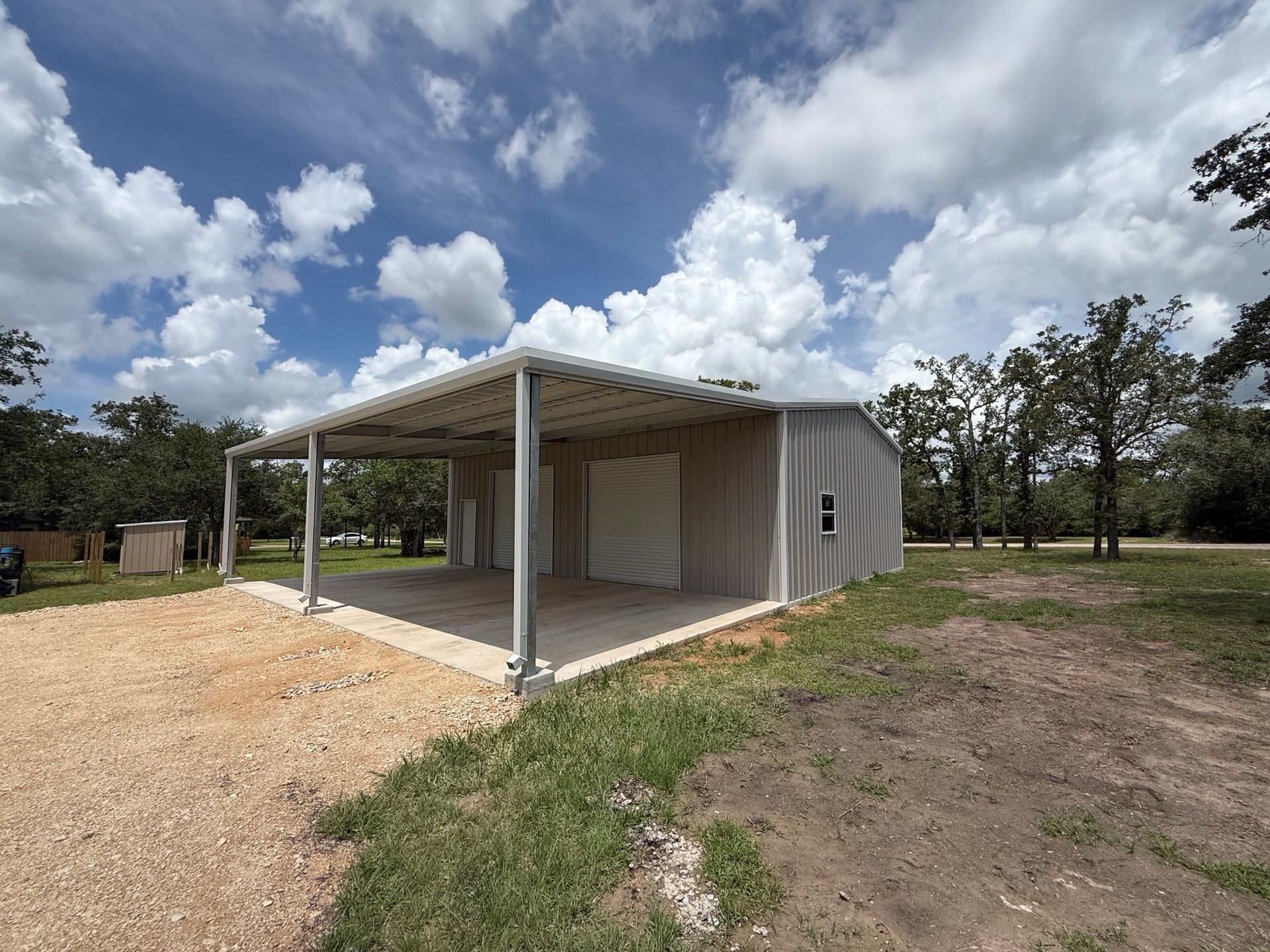 Small gray metal shed with a covered porch on a gravel lot under a cloudy sky