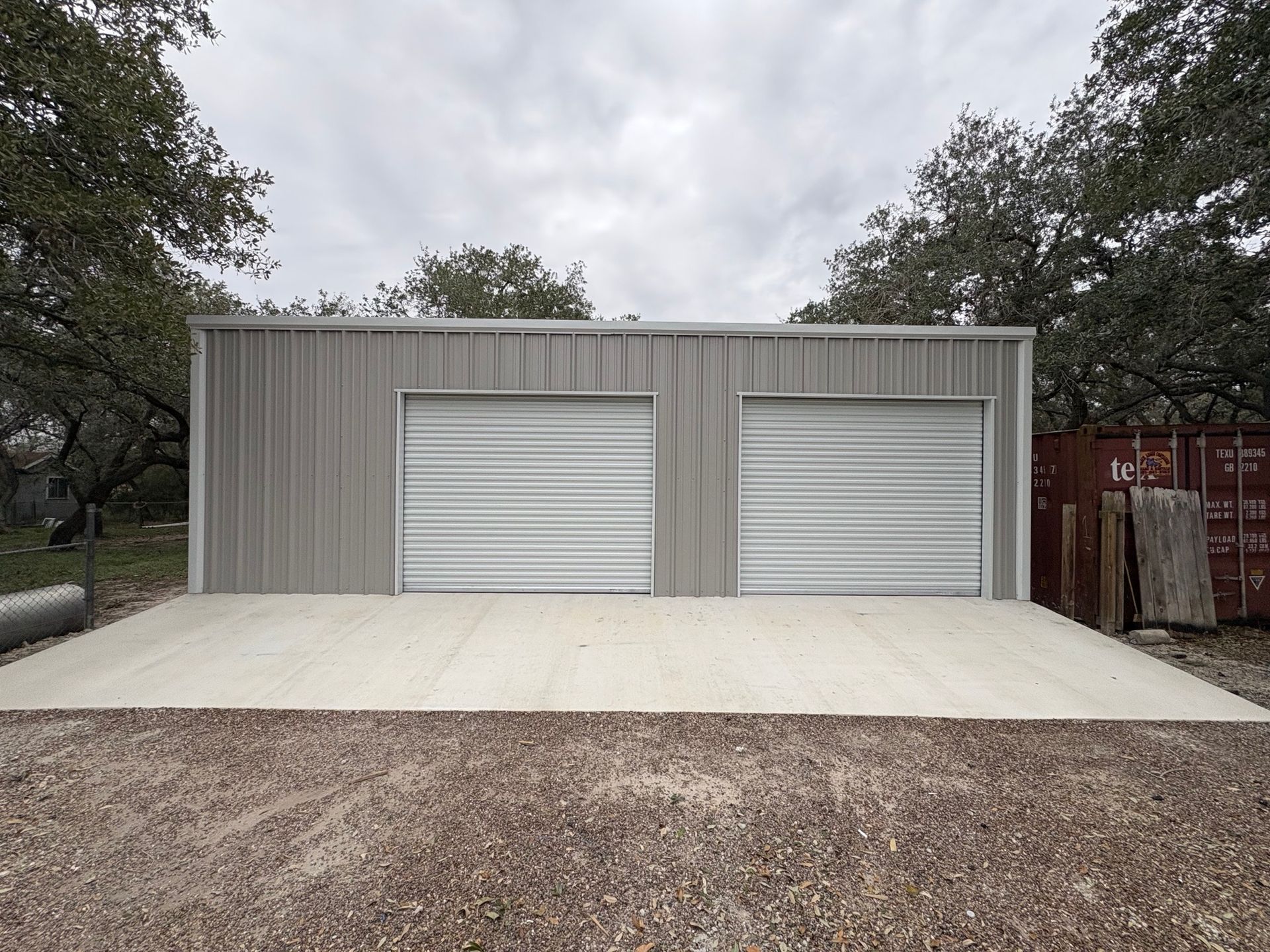 Two-car metal garage with white roll-up doors and a concrete driveway, framed by trees