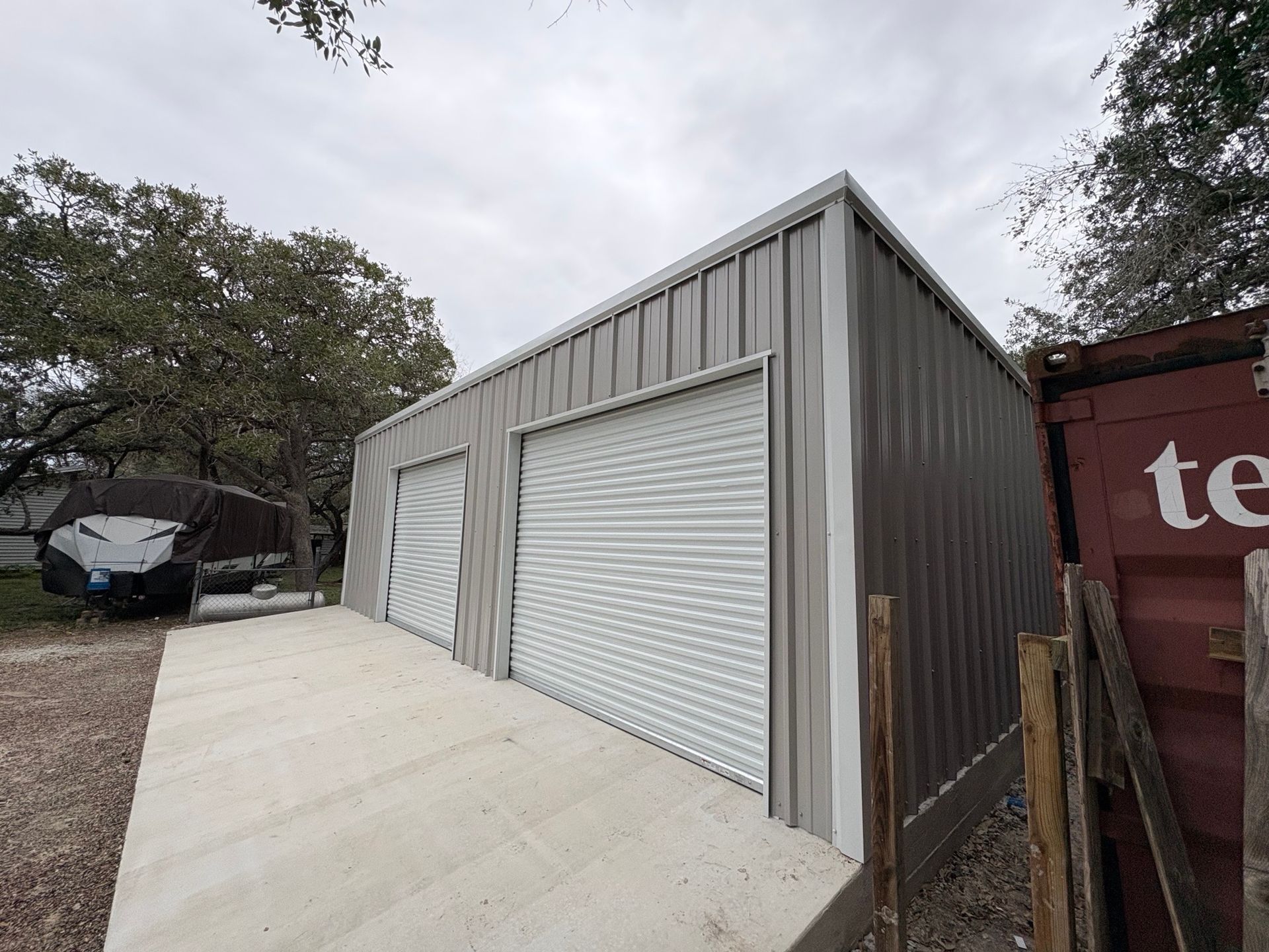 Gray metal garage with two roll-up doors beside a concrete driveway and trees outdoors