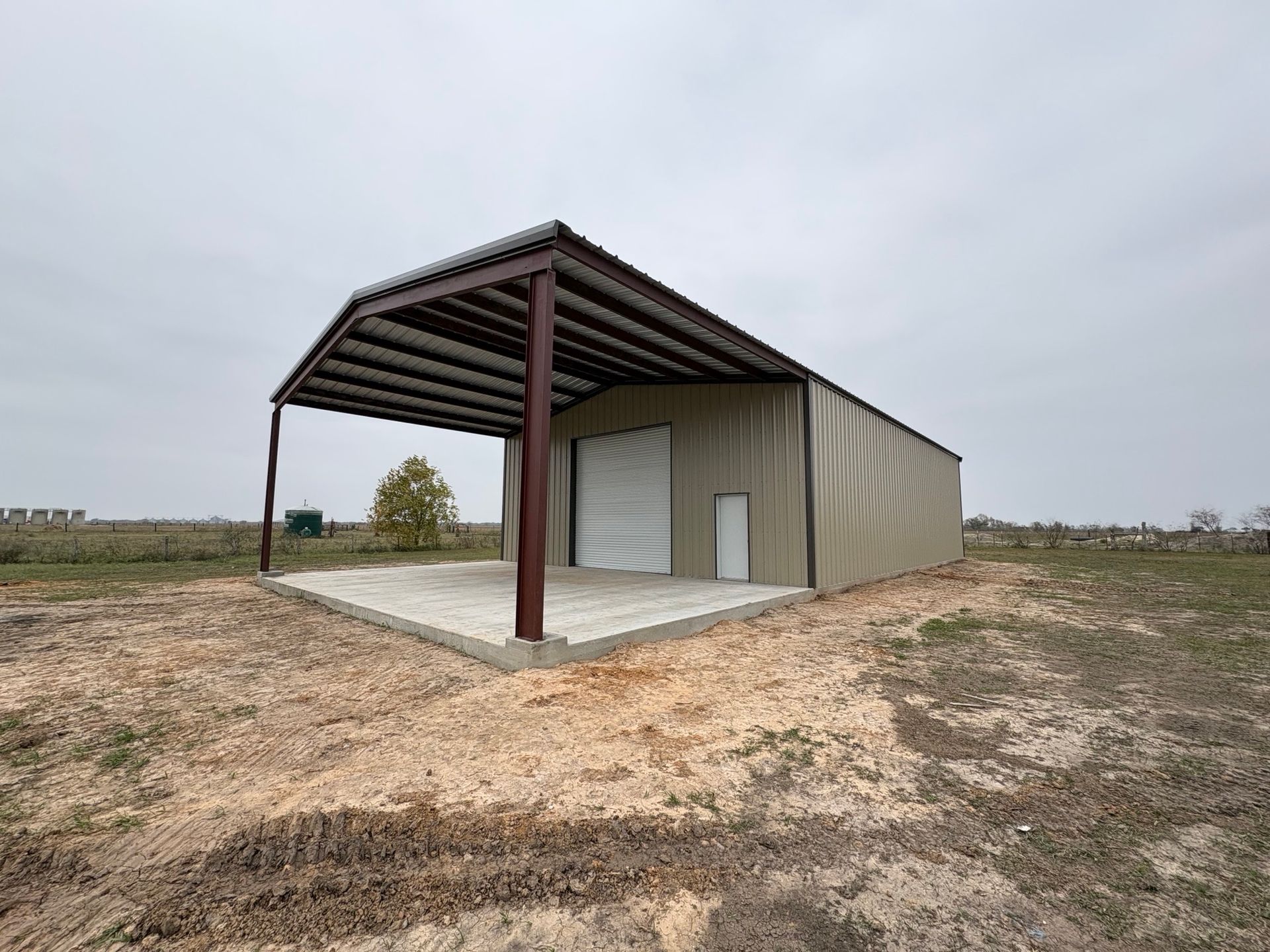 Small beige metal shed with a dark roof canopy on a concrete slab in a barren field under cloudy sky