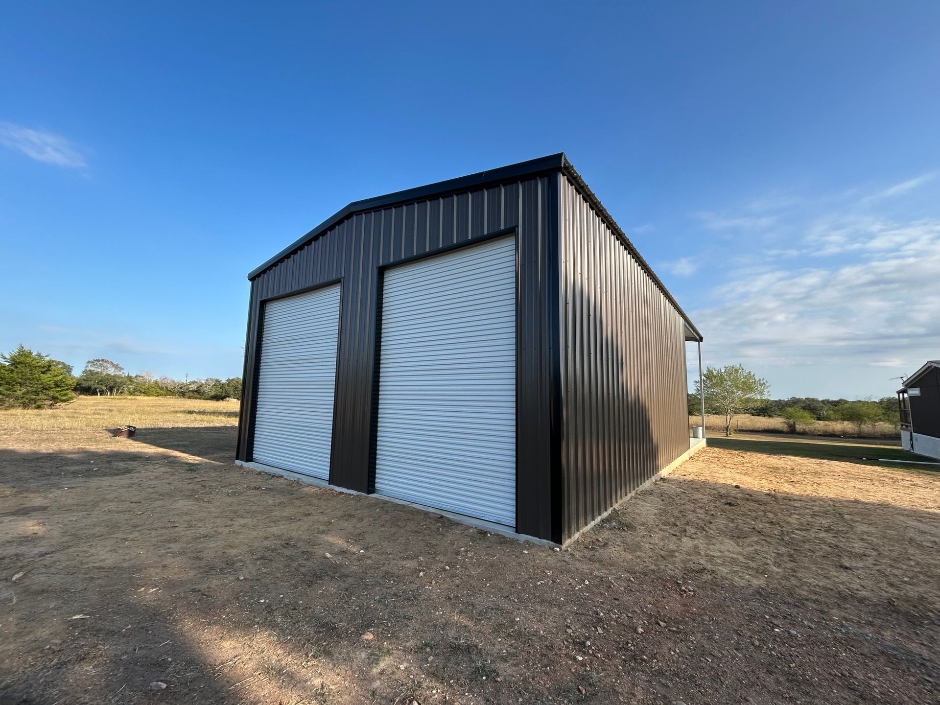 Black metal barn with two white roll-up doors on a dirt lot under a blue sky
