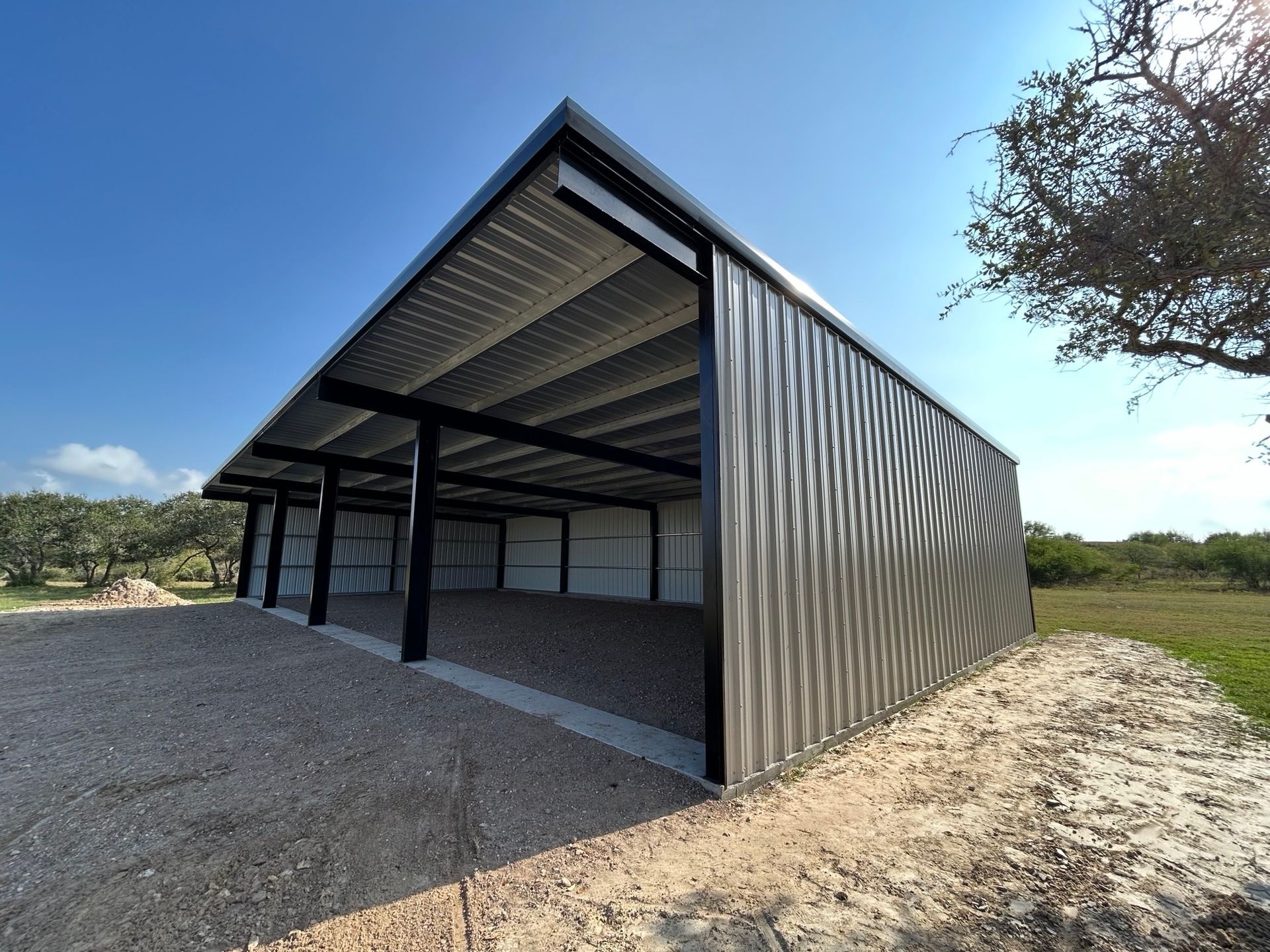 Open-sided metal shed with corrugated walls and roof on gravel in a rural field under a blue sky