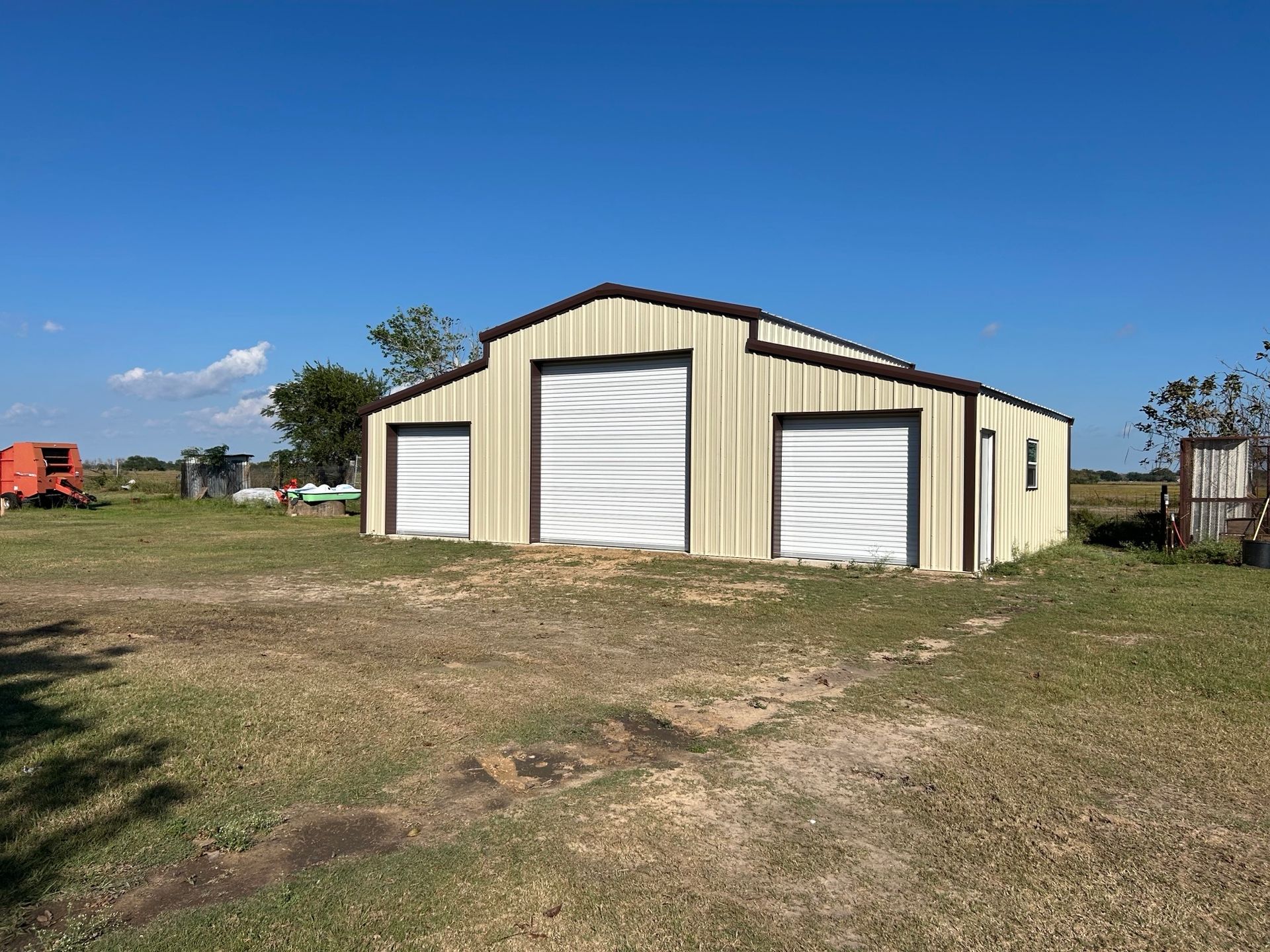 Large beige metal storage barn with white roll-up doors on a grassy lot under a clear blue sky
