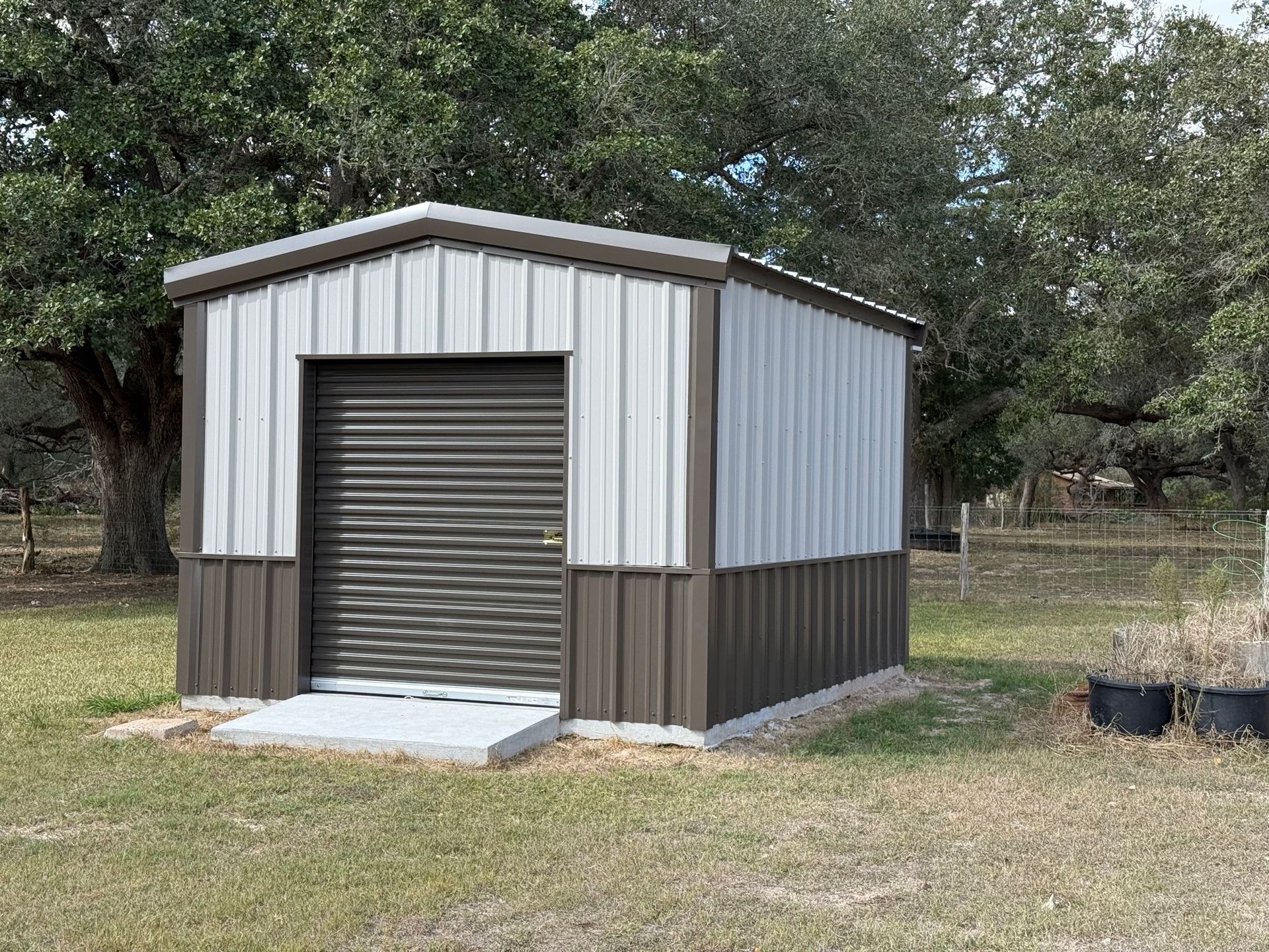 Small metal storage shed with a roll-up door and ramp in a grassy yard surrounded by trees