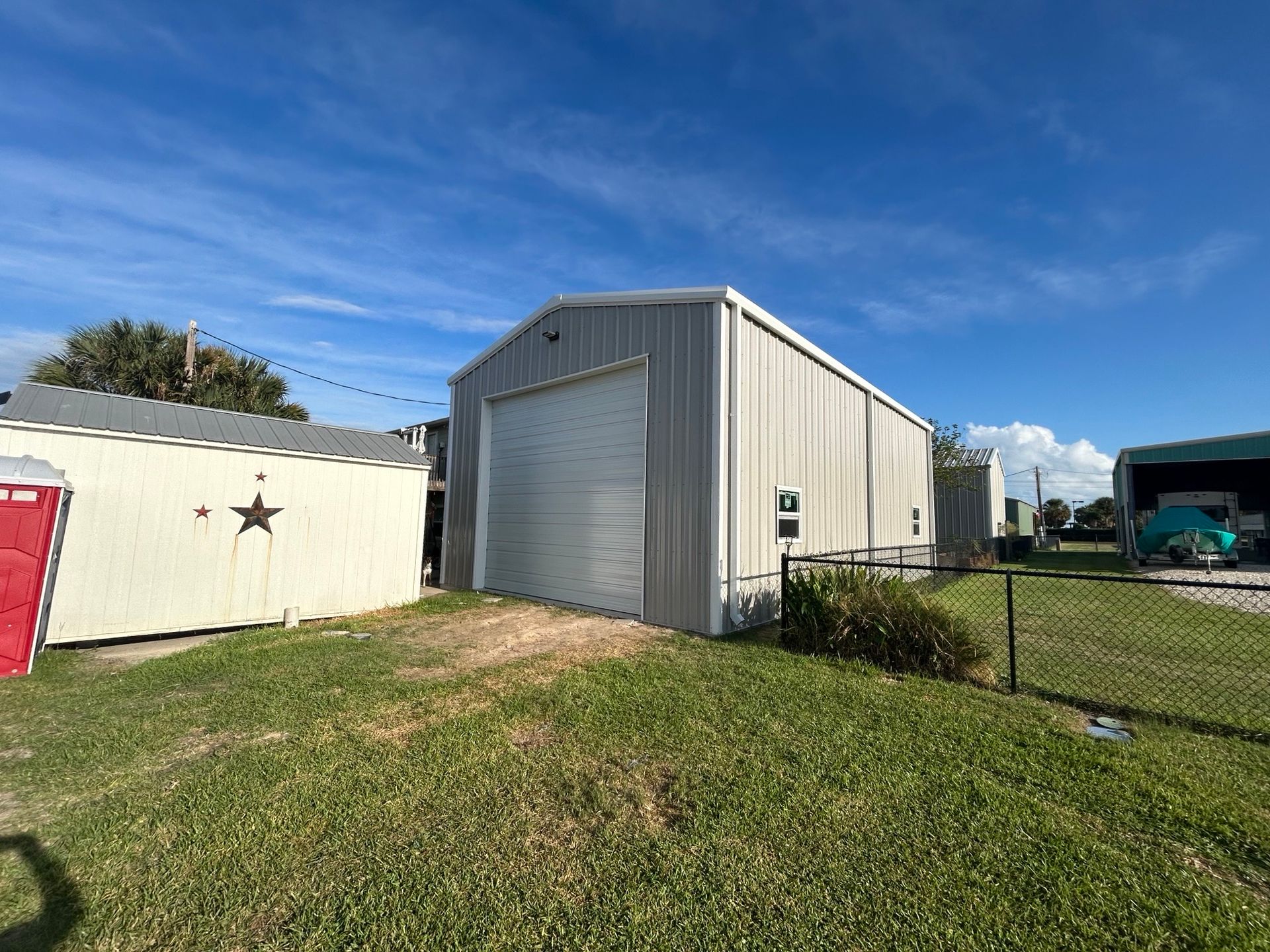 Small beige metal storage building with a roll-up door in a grassy yard under a blue sky.