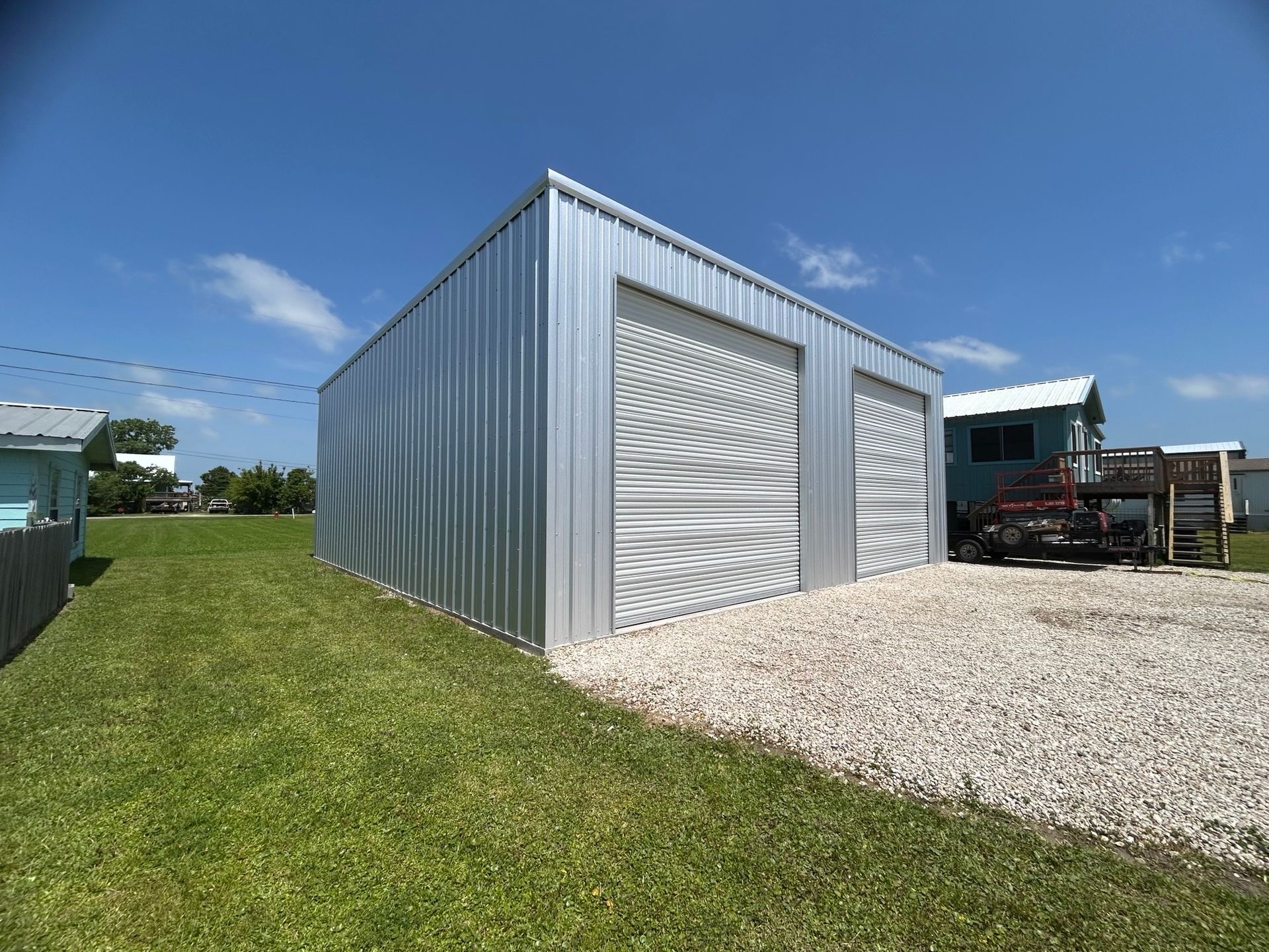 Metal storage building with roll-up doors beside a gravel drive and grassy yard under a blue sky