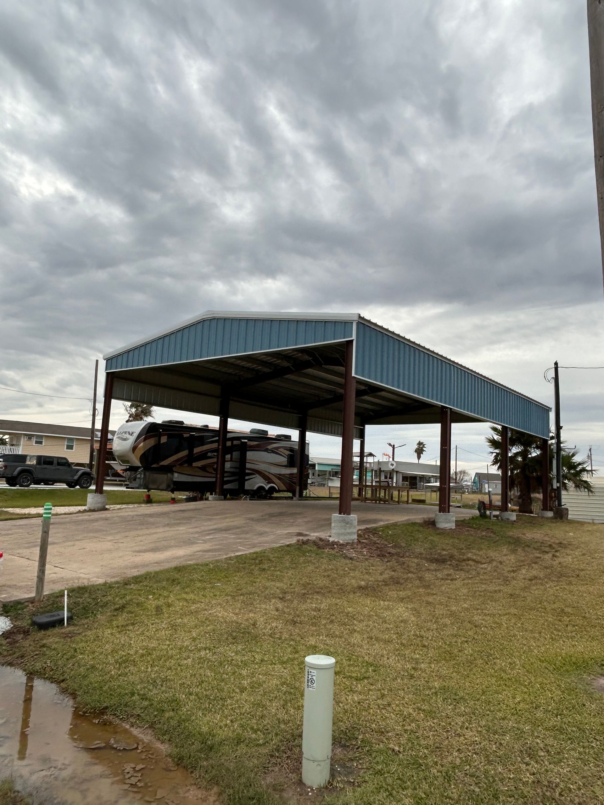 Open-sided metal shed with vehicles parked inside under a cloudy sky on a grassy lot