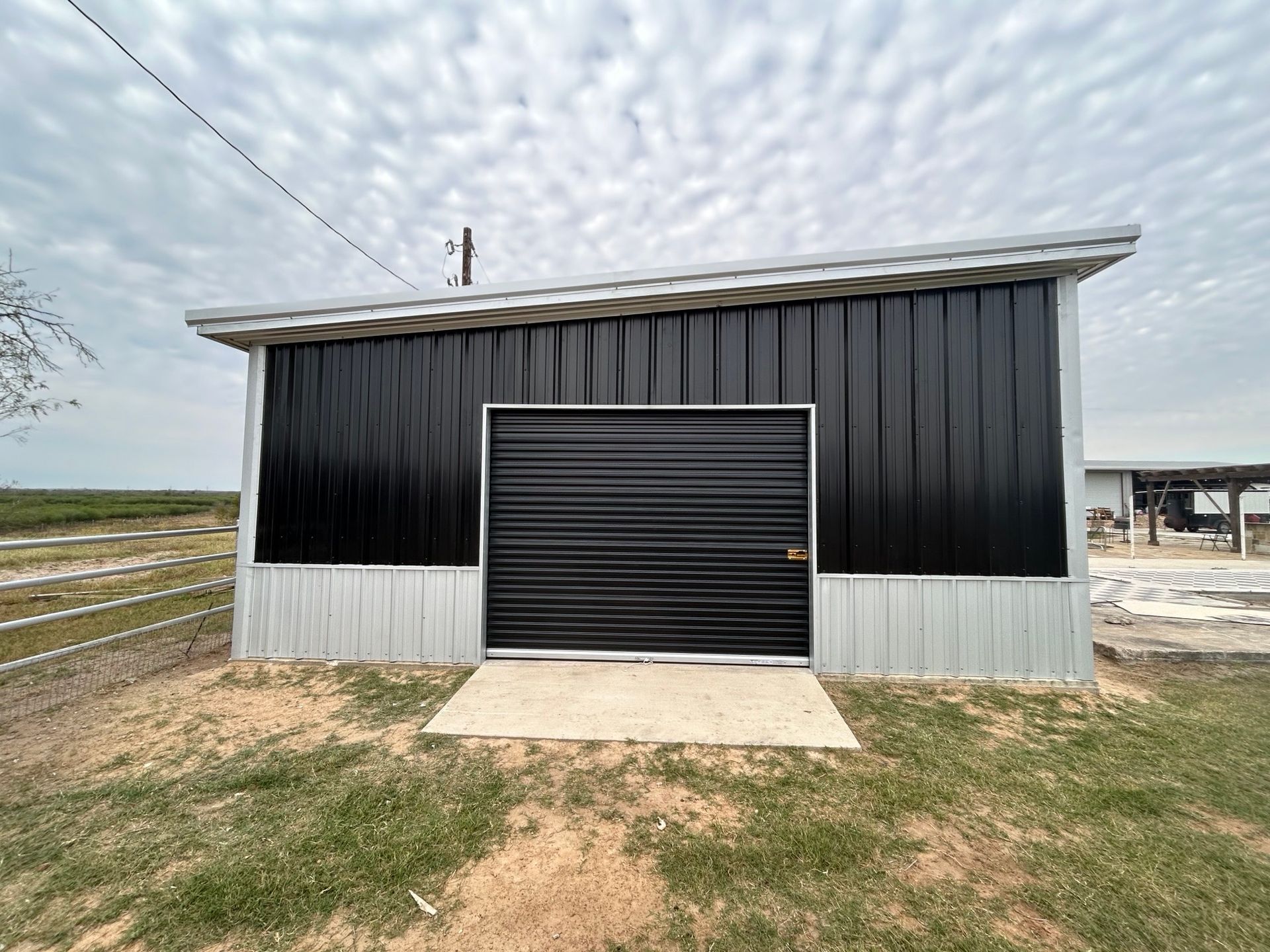 Black metal barn with a large roll-up door on a grassy rural lot under an overcast sky
