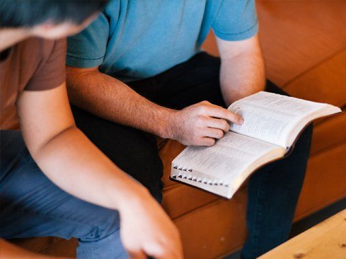 two men are sitting on a couch reading a bible .