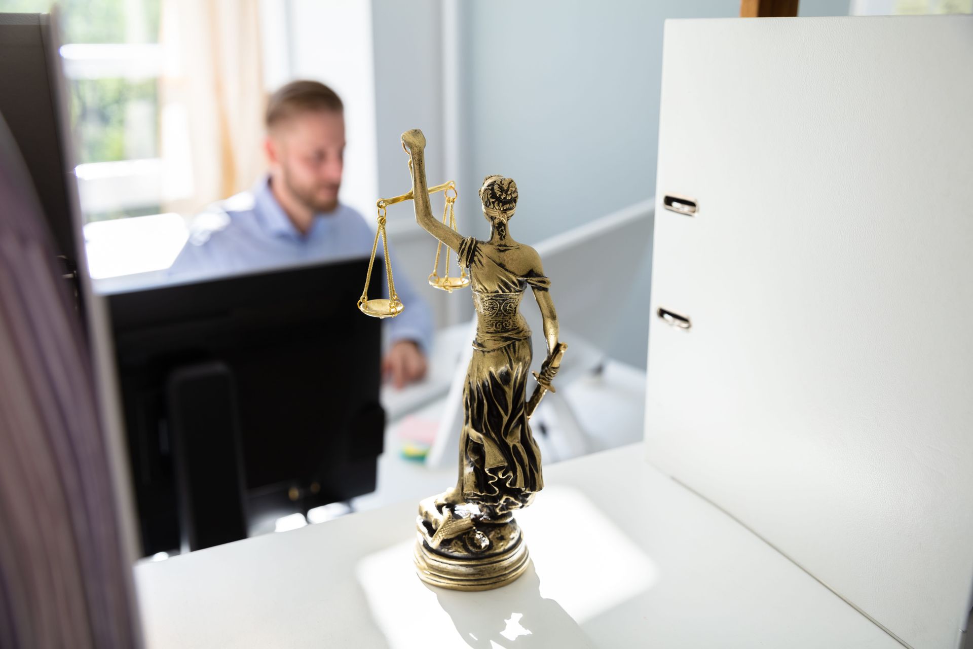 Gold Lady Justice statue on a desk in a bright office, with a blurred person in the background