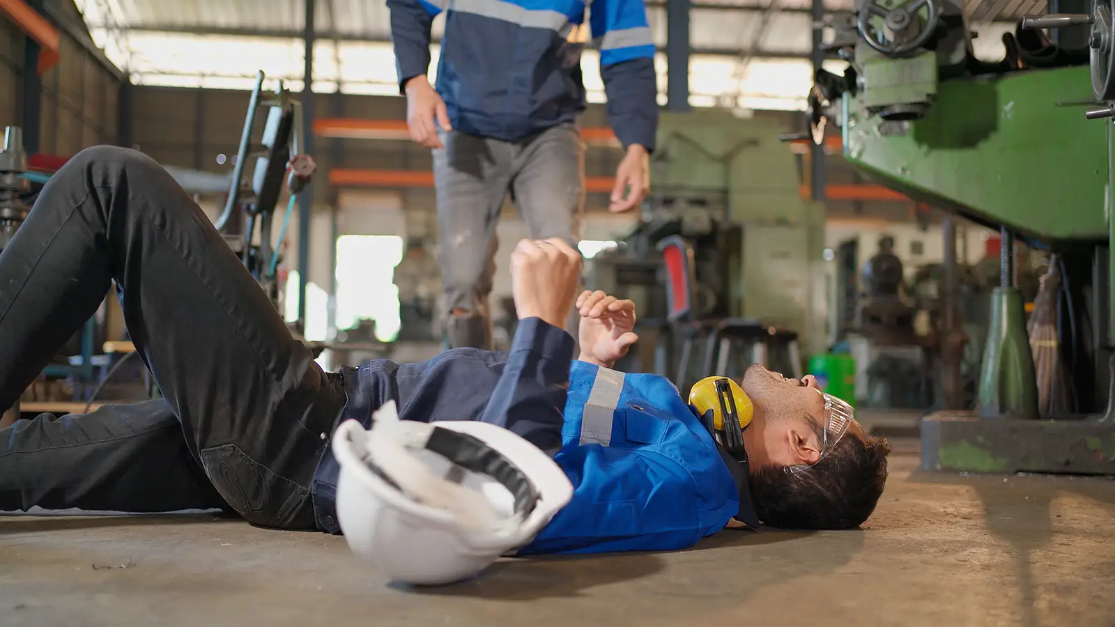 Worker lying on floor in factory, helmet nearby, another worker behind him.