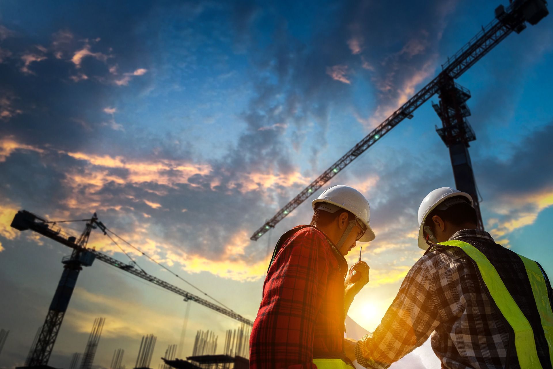 Trabajadores de la construcción mirando un plano en el sitio con grúas y puesta de sol de fondo.