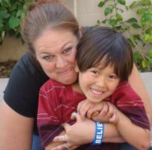 Woman embraces a smiling child; both look at the camera. Child wears a