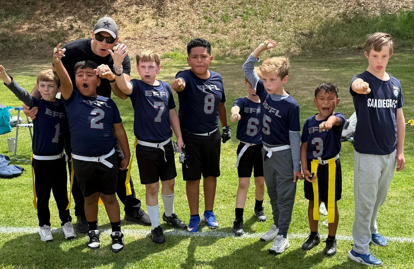 Youth football team posing on a grassy field, wearing navy jerseys and holding up number signs, with a coach behind them