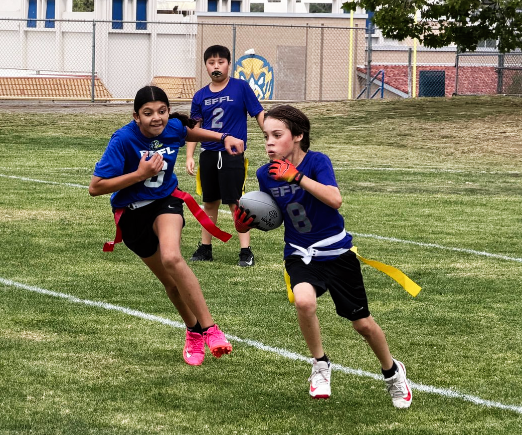 Children playing flag football on a grassy field, one in blue running with the ball while another reaches to tag him.