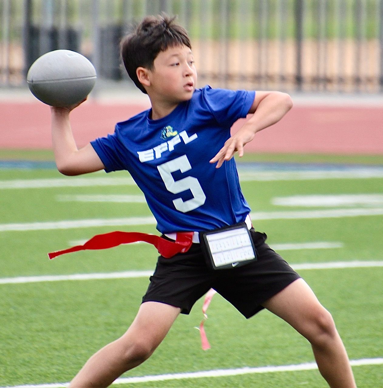 Youth football player in blue jersey #5 mid-throw on a field, holding a gray ball.