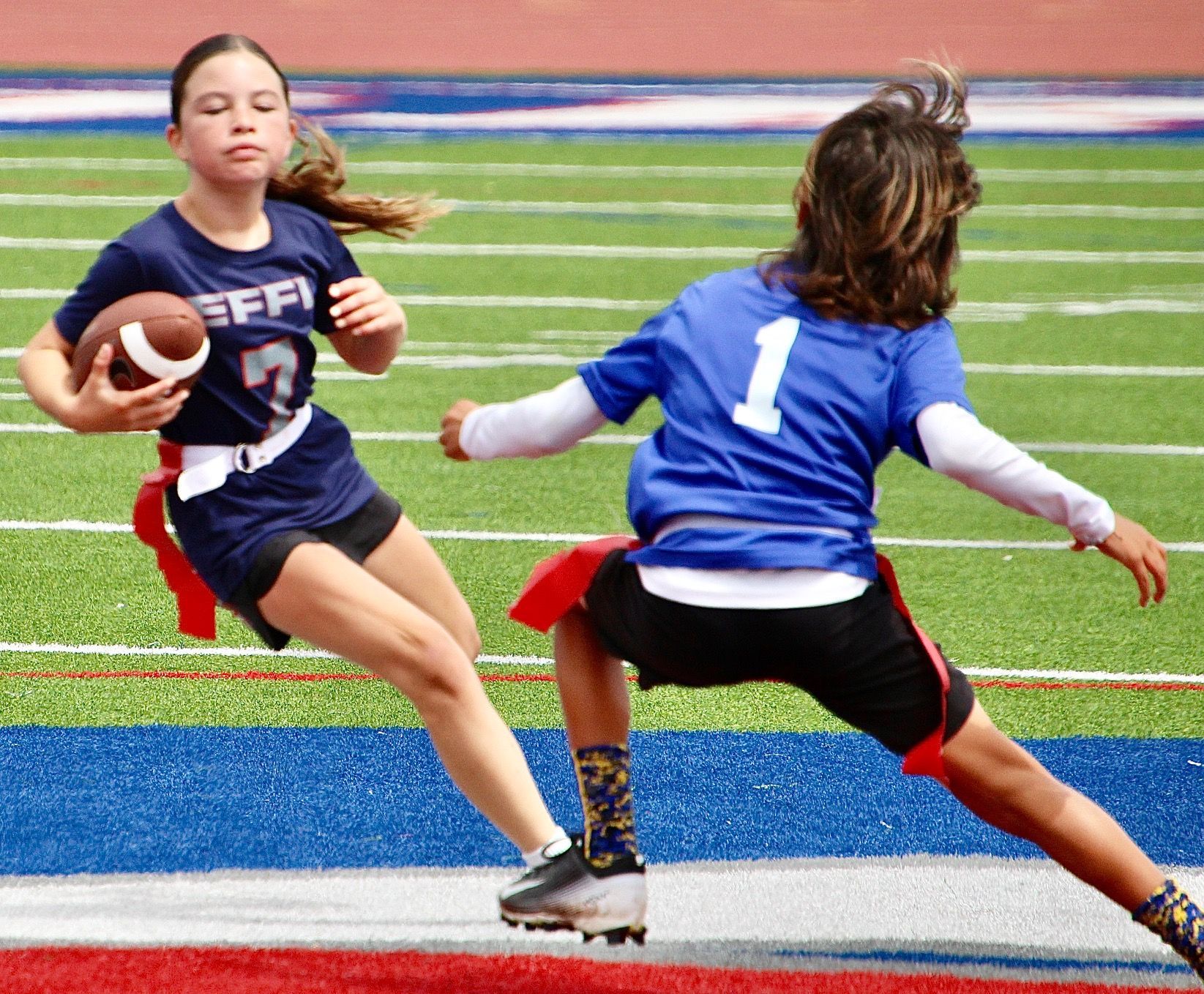 Two youth football players sprinting on a field, one carrying the ball while another in blue attempts a tackle
