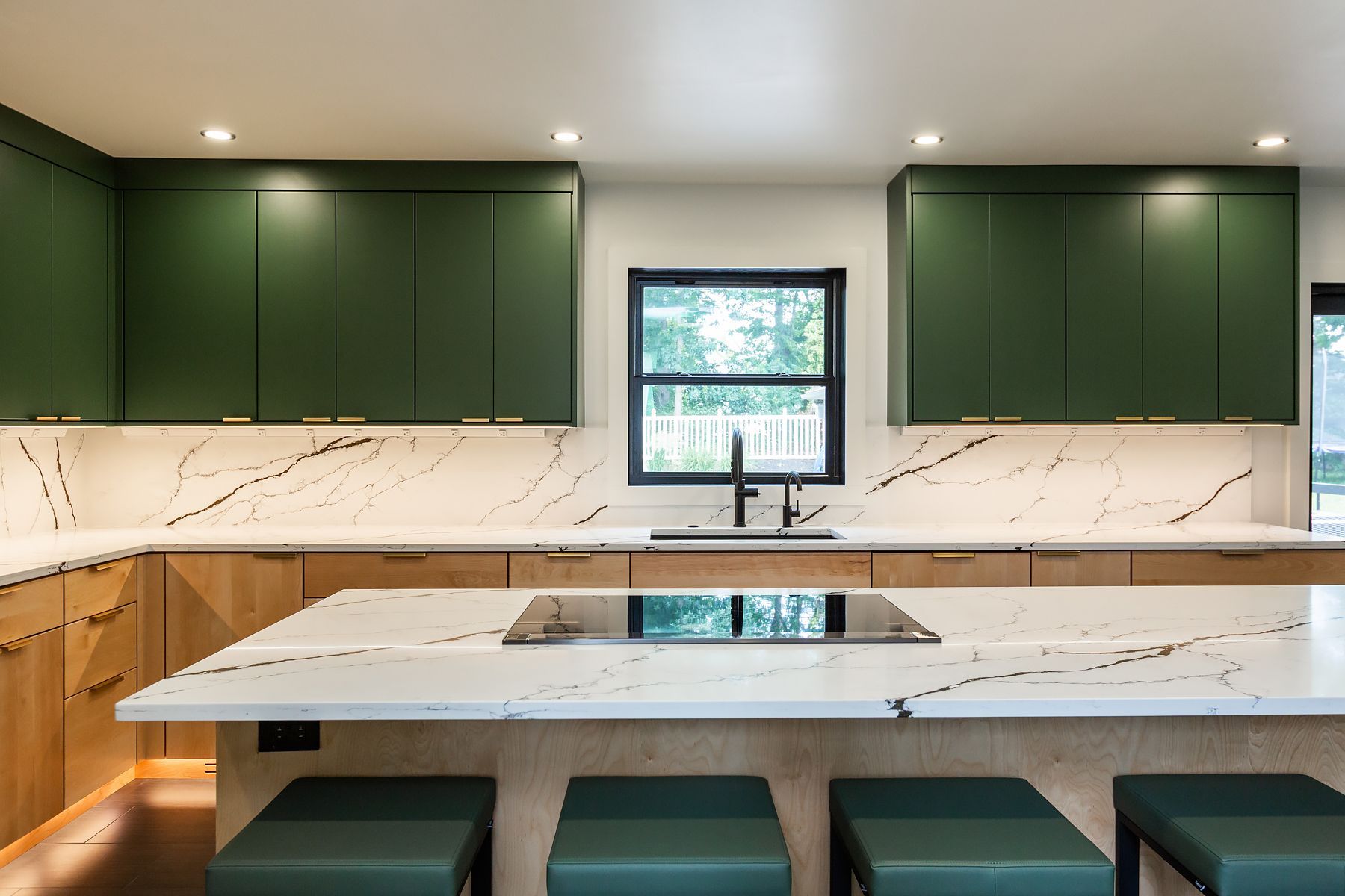 Modern kitchen with dark green cabinets, marble counters, central island, and stools by a window