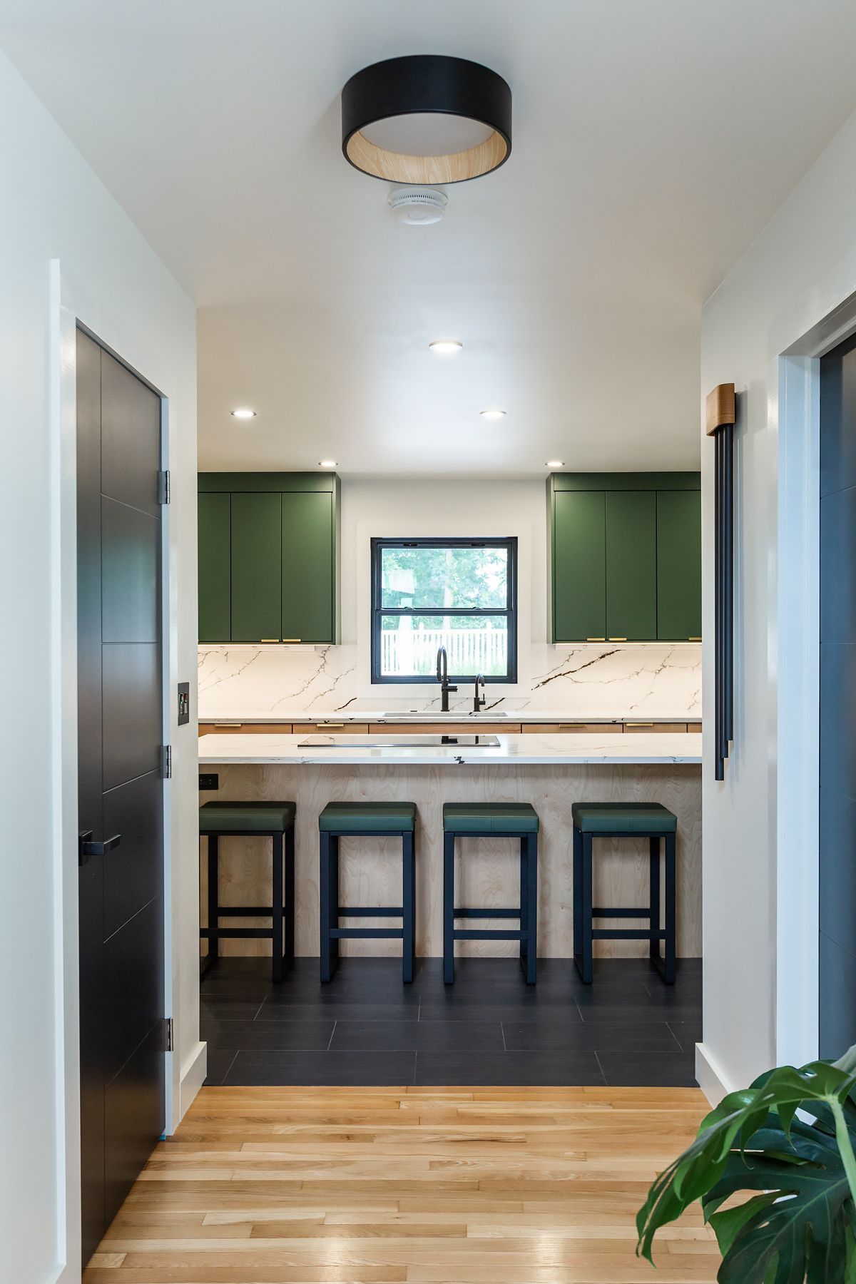 Bright modern kitchen hallway with green cabinets, marble island, and four stools centered under a window.