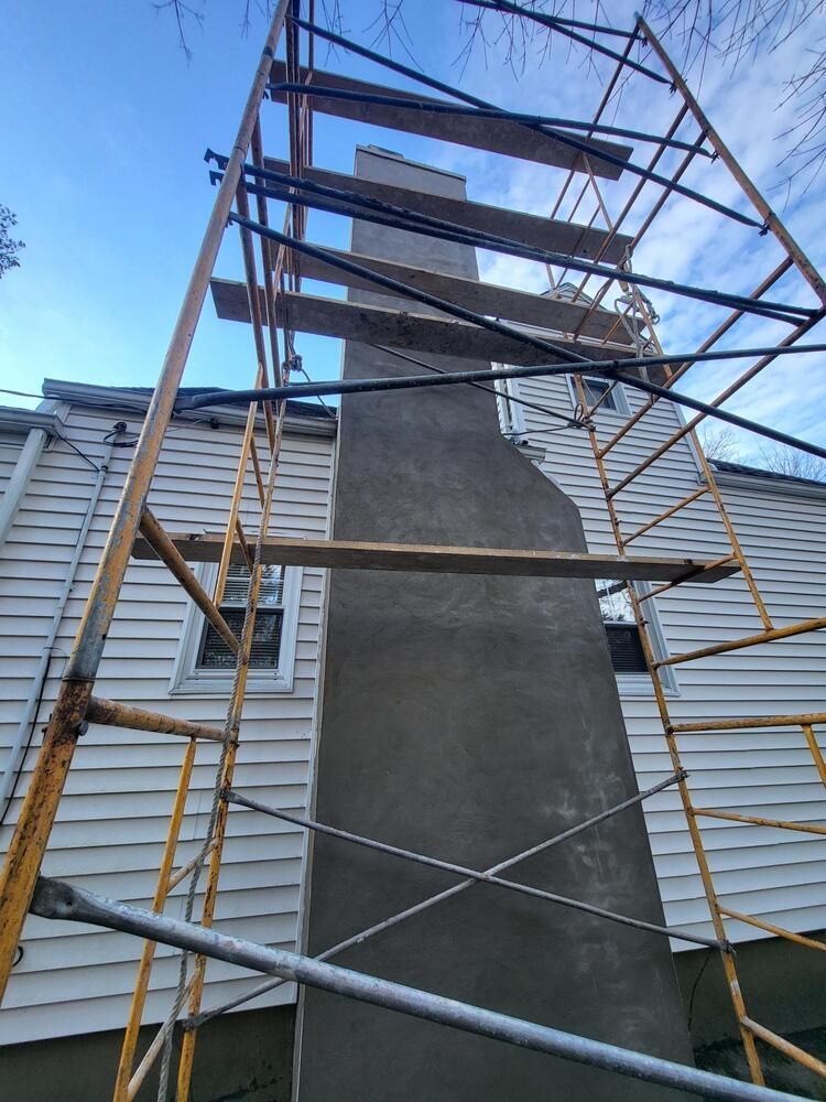 Scaffolding around a tall concrete chimney against a blue sky, with white siding on both sides