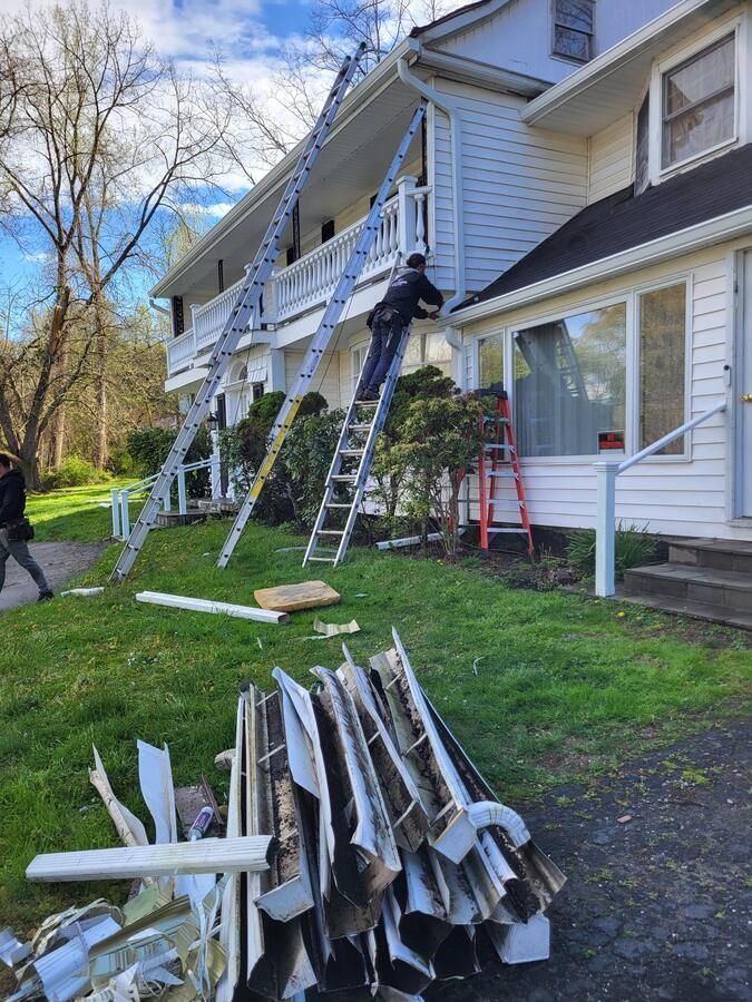 Workers on ladders repairing white siding on a townhouse, with construction materials stacked on the grass.