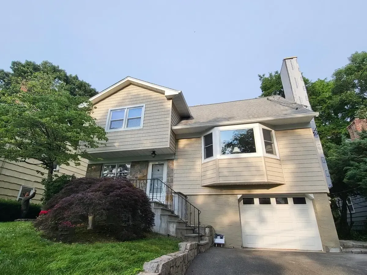 Suburban house with beige siding, white garage, and green trees on a sunny day