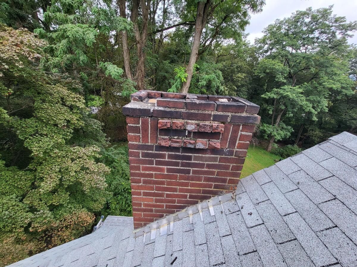 Brick chimney on a gray shingled roof beside dense green trees