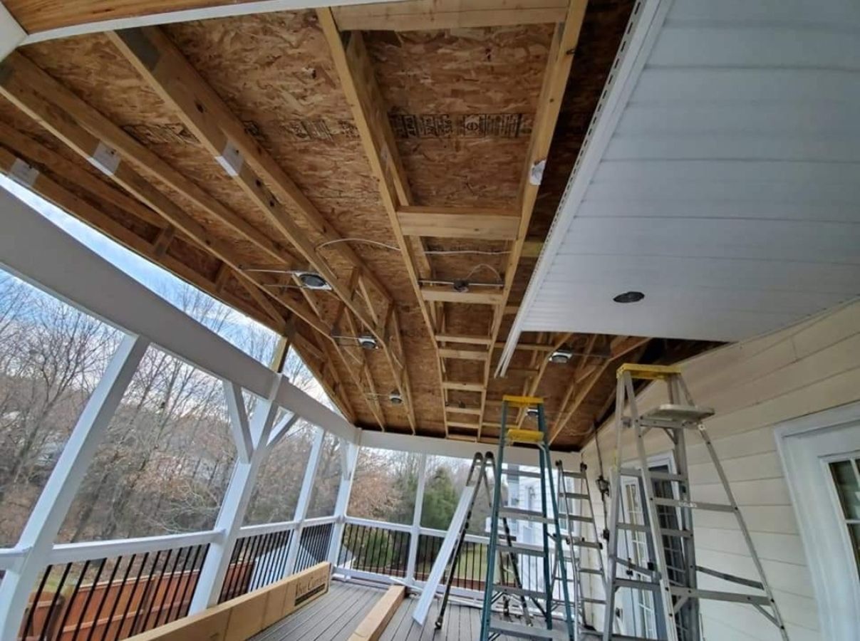 Covered porch under construction with exposed wood framing, ladders, and white siding ceiling panels