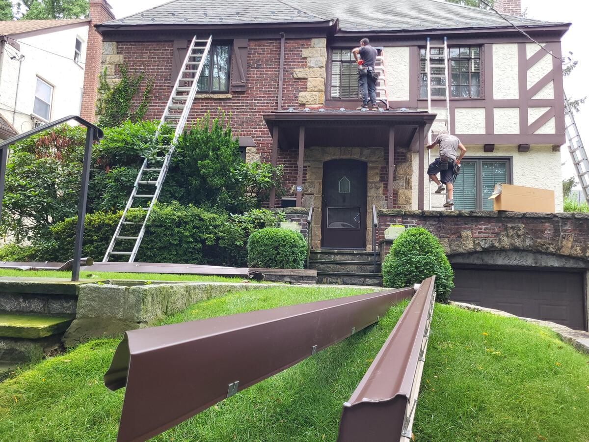 Brick house with workers on ladders and a large steel beam in the foreground on a grassy hill.