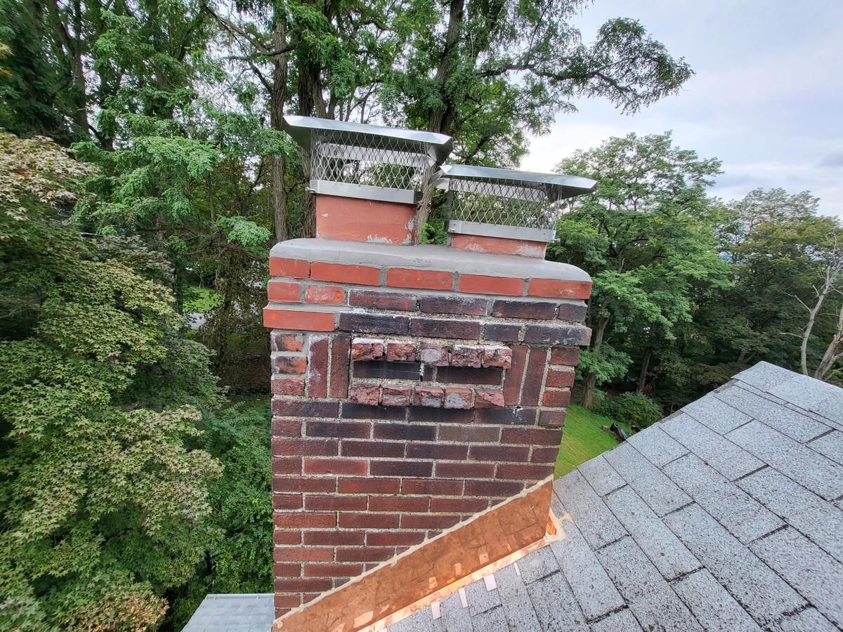 Brick chimney on a shingled roof, surrounded by green trees under a cloudy sky