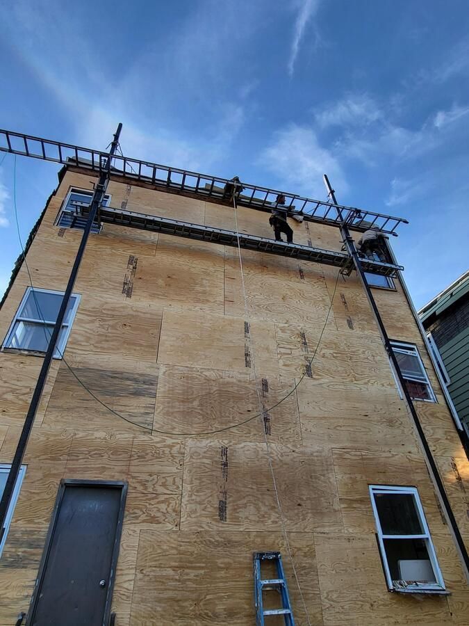 Worker on a ladder beside a tall brick building with a rooftop scaffold under a blue sky