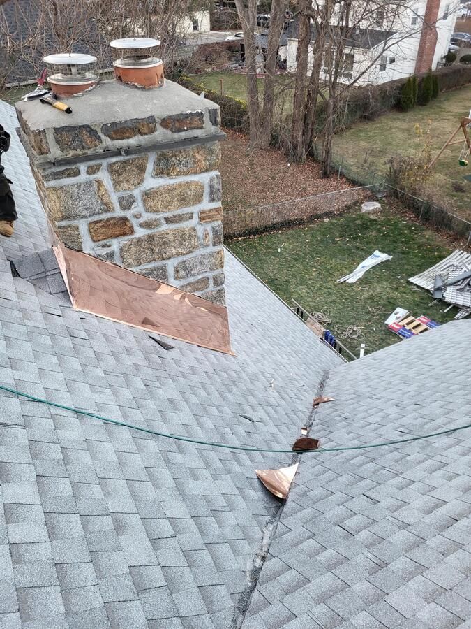 Shingled roof with a damaged seam near a stone chimney, viewed from above.