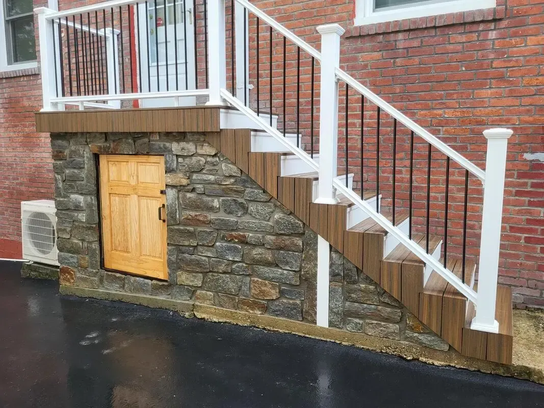 Stone basement stairs beside a brick house, with white railings and a small wooden door under the steps