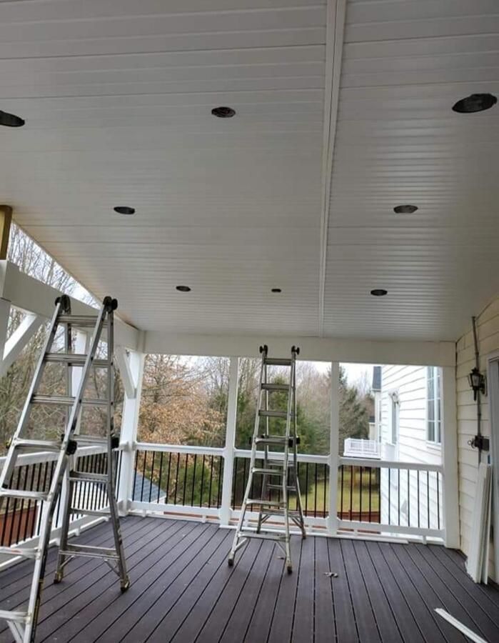 Covered porch with white ceiling and railings, two ladders, and a work stool on a dark wood floor.