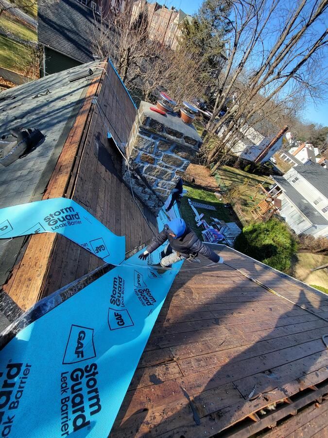Blue roofing underlayment stretched across a shingled roof beside a brick chimney and ladders.
