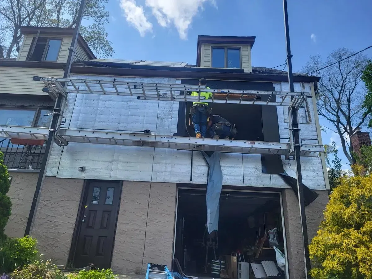 Worker on scaffolding repairing the front of a two-story house with exposed siding and roof damage